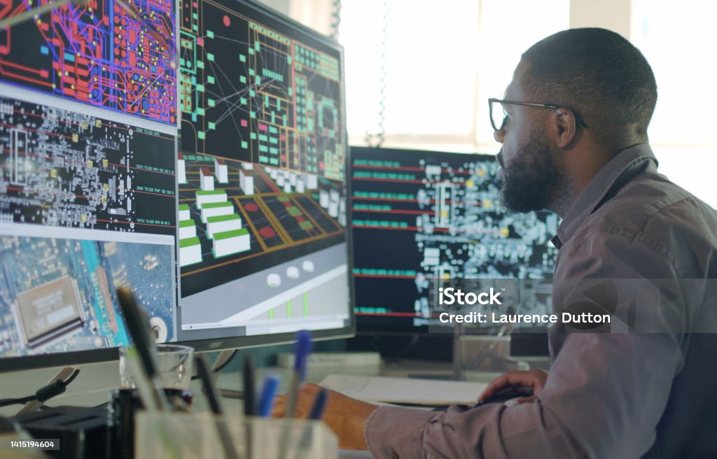 CAD electronics engineer A Stock image of an Afro-Caribbean male designing electronic circuit boards ( PCBs).
He’s sitting at a desk with a large computer screen displaying a variety of apps & PCB illustrations. Engineer Stock Photo