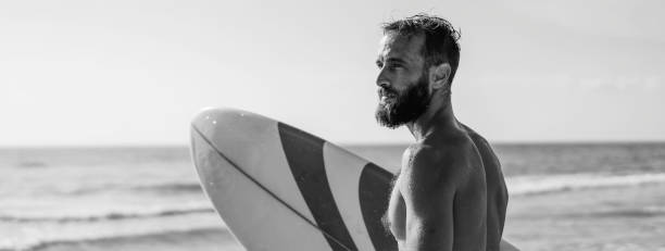 istock Horizontal banner or header with surfer holding his surfboard on the beach - Hipster man standing on the beach and waiting big waves for surfing - Fit bearded man training with surfboard to sea 1415198159