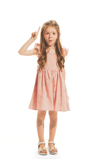 Portrait of beautiful little girl in stylish dress posing, raising finger up isolated on white studio background. Concept of emotions, facial expression, childhood, game, education. Copy space for ad