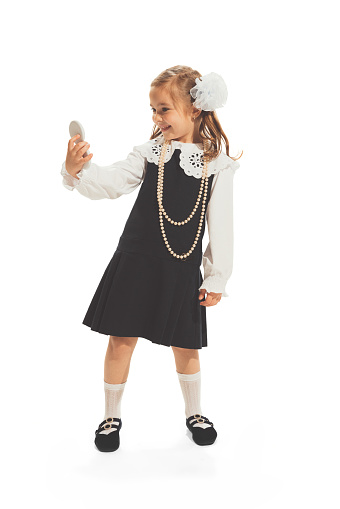 Portrait of little girl, child in school uniform with bow looking at mirror isolated over white studio background. Retro fashion. Concept of childhood, game, school, fun, education. Copy space for ad