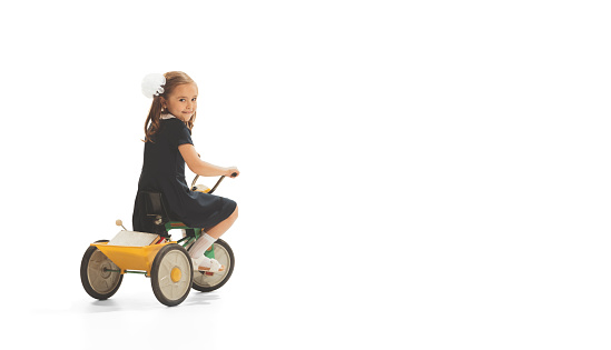 Portrait of child, girl in stylish retro dress playing, having fun, riding small bike isolated over white background. Concept of childhood, game, school, fun, education. Copy space for ad