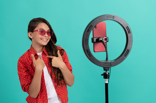 happy kid long hair in sunglasses talking on camera video blog blogging online with selfie led pointing finger on herself, vlogger.