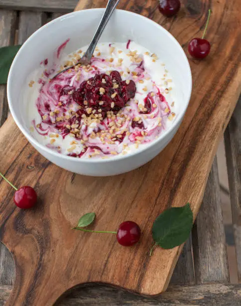 Healthy low carb breakfast, snack or dessert with a fresh greek yogurt, cooked sour cherries and roasted hazelnuts. Served in a bowl with spoon isolated on wooden table background