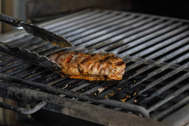 Outdoor barbecue scene shows a hand using tongs to flip several pieces of meat on a sizzling grill. Char marks, smoke, and glowing embers create a warm, savory dining moment.