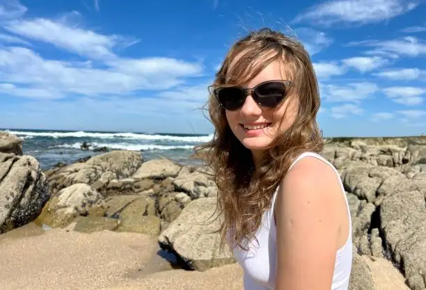 a teenage girl in sunglasses looks into the frame and smiles in the background she has the Atlantic Ocean in Portugal girl in a white T-shirt with blond hair She is a teenager 13-16 years old