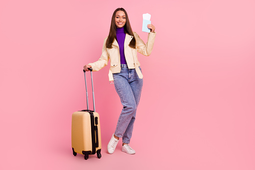 Full length photo of cute young lady with bag hold tickets wear jacket jeans boots isolated on pink color background.