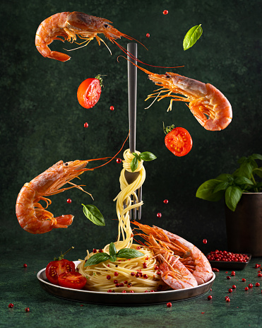 Spaghetti with red Argentine shrimps, cherry tomatoes, basil, pink peppercorn on dark green background, conceptual food levitation art photo. Seafood, copy space.