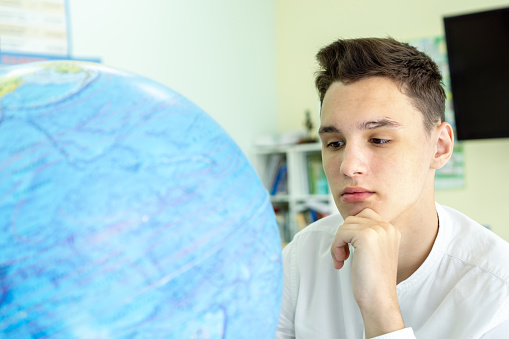 Portrait of a thoughtful young man in a white shirt sitting at a table in front of a globe. The concept of geography lesson, travel