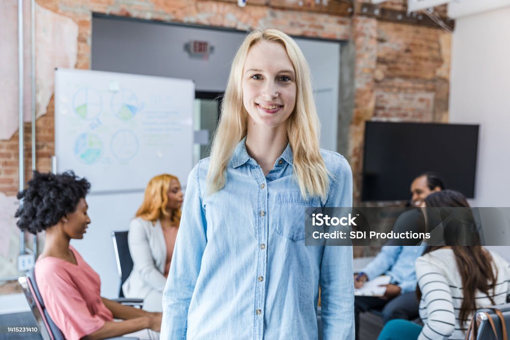 Young woman smiles for camera while colleagues continue discussion As her colleagues continue their discussion, the young adult woman smiles for the camera. 20-24 Years Stock Photo