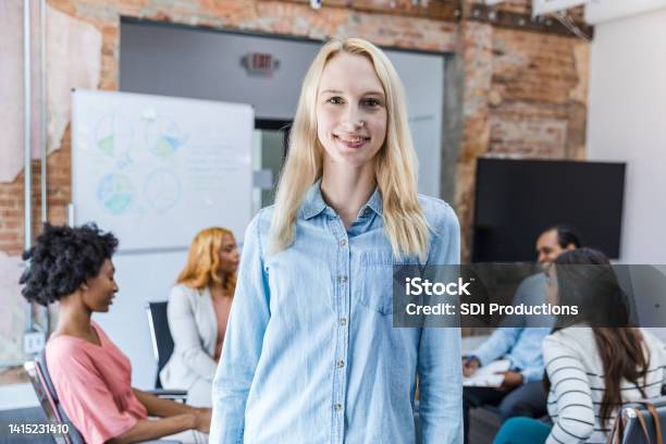 Young Woman Smiles For Camera While Colleagues Continue Discussion Stock Photo - Download Image Now