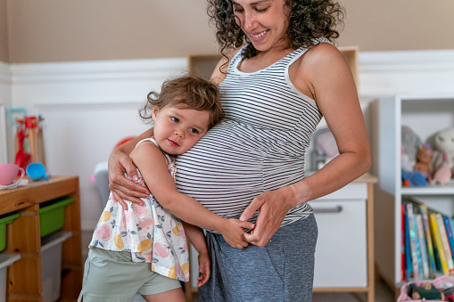 A pregnant thirty two year old ethnic woman is kneeling on the floor of a play room and interacting with her toddler daughter. The mother and daughter are happy as the girl affectionally holds her moms pregnant belly and smiles directly towards the camera.