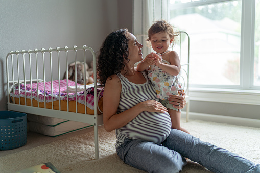 A thirty something year old ethnic woman is interacting with her toddler daughter. They are sitting on the floor while she holds her toddler on her hip and has one hand under her pregnant belly.