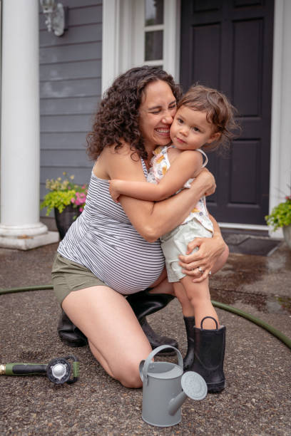 A pregnant Mama and her toddler are hugging while taking a break from watering plants together outside