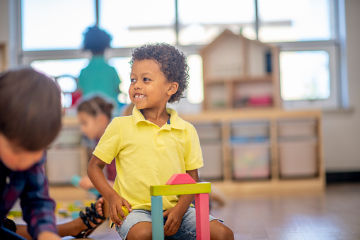 A young African boy sits on the floor among his Kindergarten peers building with colorful blocks. He is dressed casually and looking across the classroom and smiling as he focuses on his task.