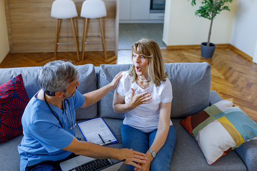 A mature Caucasian female patient and a mature Caucasian male doctor are enjoying a discussion together, while sitting on her living room sofa, during a check-up.