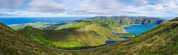 Wide panorama from viewpoint Miradouro de pico da Barrosa. Panoramic landscape with beautiful blue crater lake Lagoa do Fogo in Natural Reserve with green forest, mountains and hills, sea coast and asphalt road. Sao Miguel, Azores, Portugal. Sunny day.