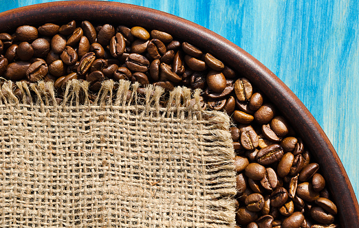 Roasted coffee beans in a ceramic plate on a turquoise background. A piece of burlap for text. Grains of a tropical plant.