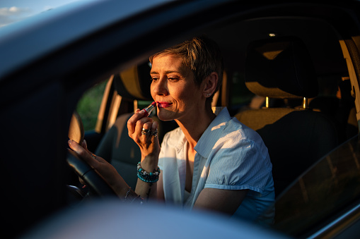 While driving in a car, a mature woman takes the opportunity to apply her make-up by looking at herself in the mirror.