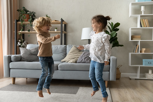 Happy adorable small children siblings dancing to disco pop music on floor carpet in modern living room. Joyful cute little brother and sister or friends having fun together at home, hobby activity.