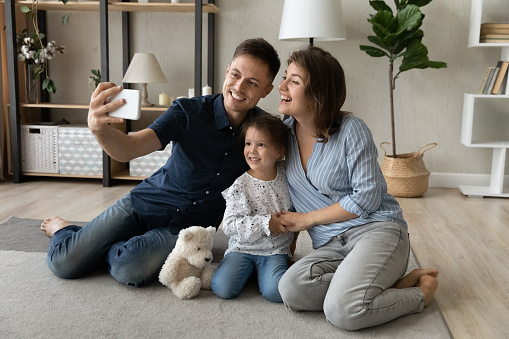Happy affectionate young couple parents and small adorable kid daughter posing for selfie photo or recording video for social network sitting together on floor carpet in stylish modern living room.