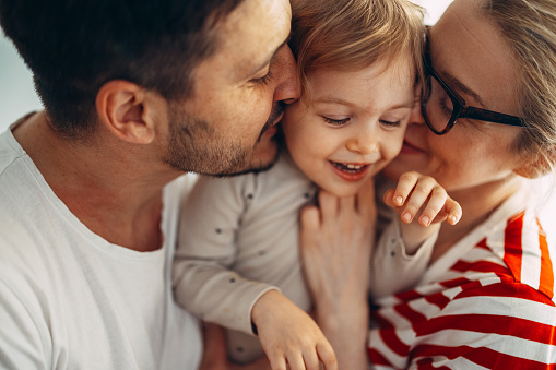 Close up shot of a cheerful couple hugging and kissing their smiling daughter in the middle.