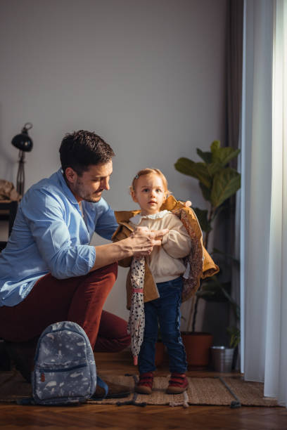 Happy Dad Getting His Daughter Ready to Go Outside Handsome cheerful dad is ready to go to work and he is crouching next to his daughter preparing her to go outside too. There is a small backpack on the floor. He is dressing her in a yellow jacket. girls getting ready to go out stock pictures, royalty-free photos & images