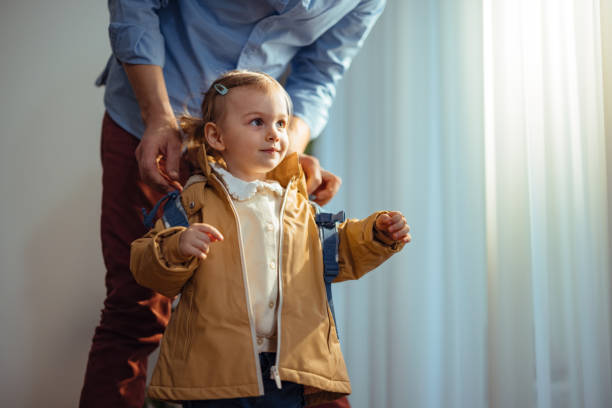 Anonymous Dad Getting His Daughter Ready to Go Outside Unrecognizable father is dressed up for work and he is standing behind his daughter preparing her to go outside with him. He is putting on her small backpack while she is looking away. girls getting ready to go out stock pictures, royalty-free photos & images