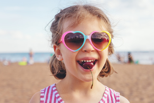 happy laughing girl 5 years old in heart-shaped sunglasses and with cherries in your mouth on beach in summer day