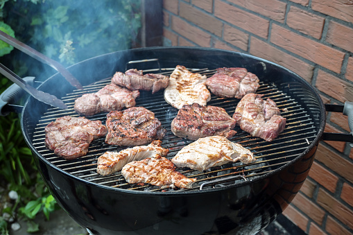 Steaks of various meats on a mobile round grill are roasted over charcoal at a barbecue party in the backyard, outdoor cooking concept, copy space, selected focus, narrow depth of field