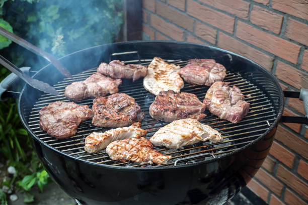 Steaks of various meats on a mobile round grill are roasted over charcoal at a barbecue party in the backyard, outdoor cooking concept, copy space, selected focus stock photo