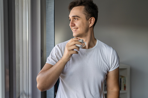 One man young adult male caucasian with brown hair standing by the window at home in morning day confident looking to the side happy smile confident wear white shirt with cup of coffee copy space