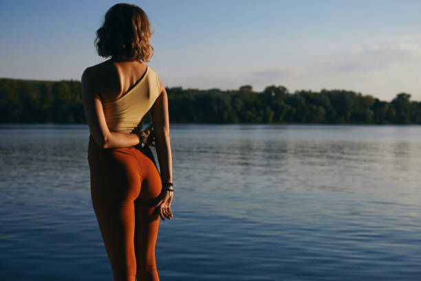 Back view of a yogi woman is looking at the river while standing on the shore at dusk. Back view of a yogi woman is looking at the river while standing on the shore at dusk. yoga pants stock pictures, royalty-free photos & images