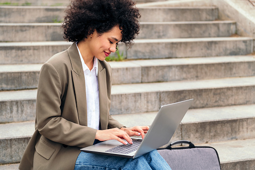 smiling young woman working with her laptop computer sitting on a city staircase, concept of business and urban lifestyle, copy space for text