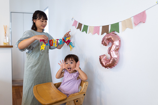 Mother setting up birthday decoration, charming daughter sitting on chair