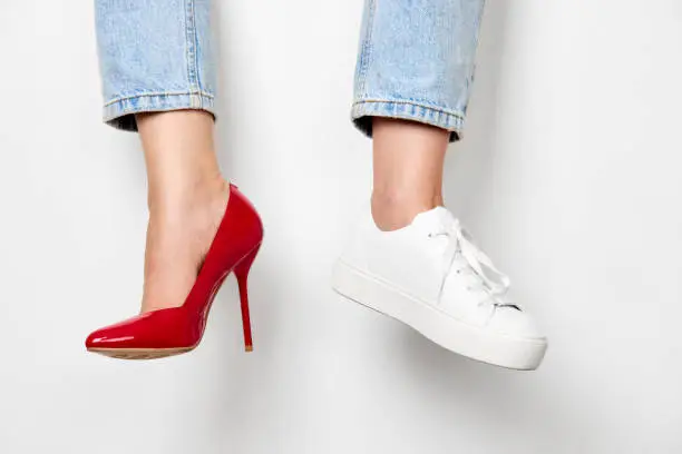 female legs in white sneakers and red high heels shoes on white background. choice between comfortable and beautiful shoes