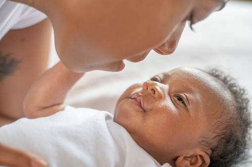 An infant African boy lays on a bed looking up at his mother. The young mother is leaning in close to his face as she looks down at him tenderly. They are both dressed casually.