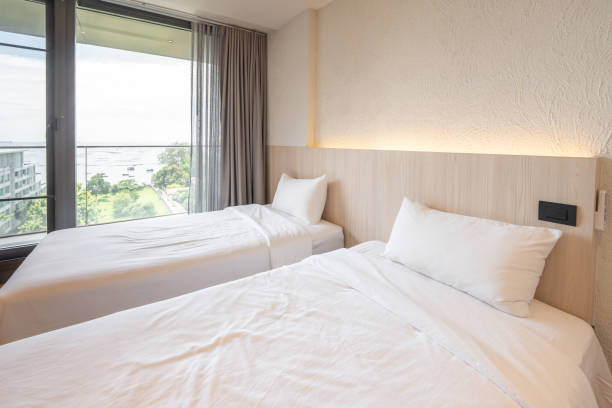2 lits simples avec literie blanche sont dans la chambre crème de l’hôtel qui allume la lumière de la tête de lit et ouvre le rideau gris. - curtain balcony bed sheet photos et images de collection