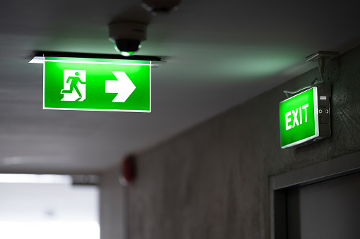 Una señal de salida de incendio verde se coloca en el techo a lo largo del pasillo poco iluminado y hay una señal de salida verde en la puerta de salida. photo