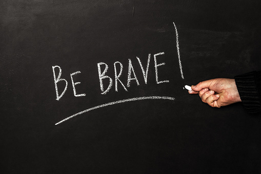 A man writes on a black board with white chalk the phrase Be brave!