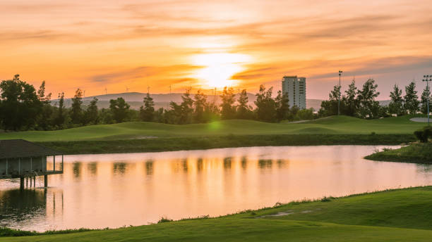 Orange red dawn sun above horizon, close-up lake pond foreground water texture surface calm reflection, trees high building background park city, hills green grass mountains, fabulously mysterious. sol rojo anaranjado del amanecer sobre el horizonte, estanque del lago de cerca en primer plano textura del agua superficie reflexión tranquila, árboles altos edificio fondo parque ciudad, colinas hierba verde montañas, fabulosamente misterioso