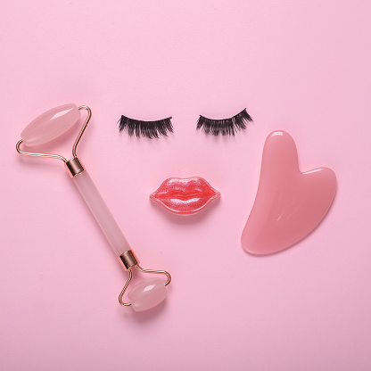 False eyelashes with lips in the shape of a face and facial massage roller on pink background. Beauty minimal still life. Flat lay