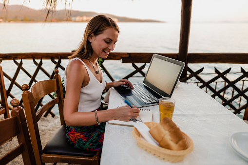 Smiling young woman sitting in a restaurant at the seaside, using a laptop and doing homework during summer vacation