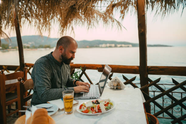 istock Bearded man using a laptop in a restaurant at the seaside 1415313310