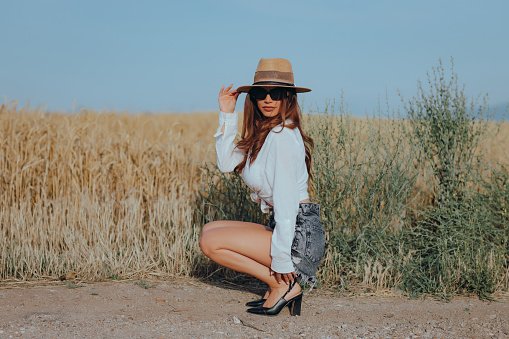 Fashionable young woman wearing a hat, denim shorts and a white shirt enjoying a sunny summer day in nature