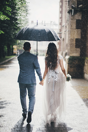 Rain drops on background of stylish bride and groom walking under umbrella and holding hands at old church in rain. Beautiful wedding couple walking together in rainy street.
