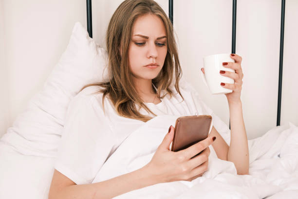 istock Young, serious, focused woman using her phone while lying in bed in the morning with cup in her hand. Awakening. Girl reads bad news, social networks, online communication. Morning routine. 1415317355
