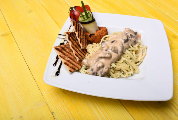 Main course concept. Pasta with white sauce and decor on yellow wooden background. Table served for one in restaurant or cafe. Italian spaghetti with chicken and mushrooms on square plate.