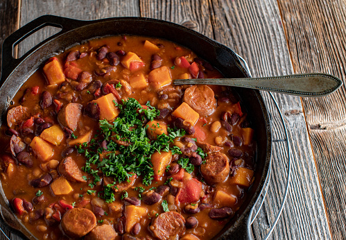 Spicy mexican or tex mex stew with chorizo sausage, kidney beans, sweet potatoes and vegetables. Served in a rustic cast iron pan isolated on wooden background.