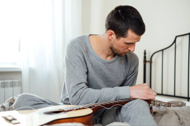 Young handsome man changing string on acoustic guitar while sitting on bed at home