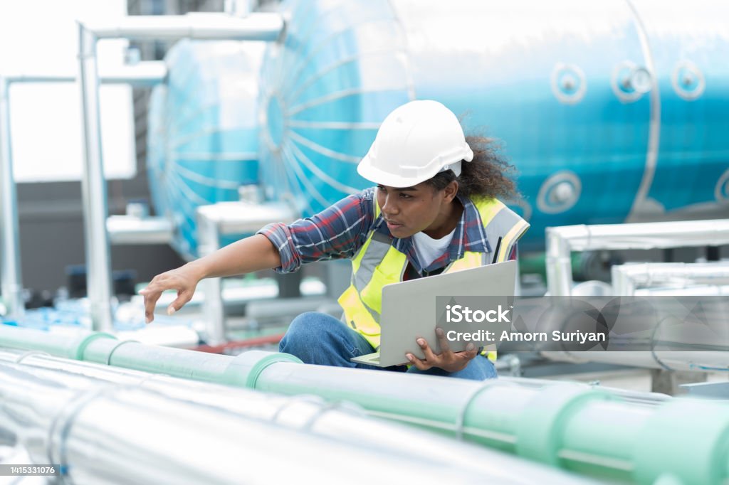 Female engineer working with laptop computer for checks or maintenance in sewer pipes area at construction site. African American woman engineer working in sewer pipes area at rooftop of building Water Pipe Stock Photo